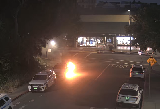 Early morning street scene with several parked cars, one of which is on fire